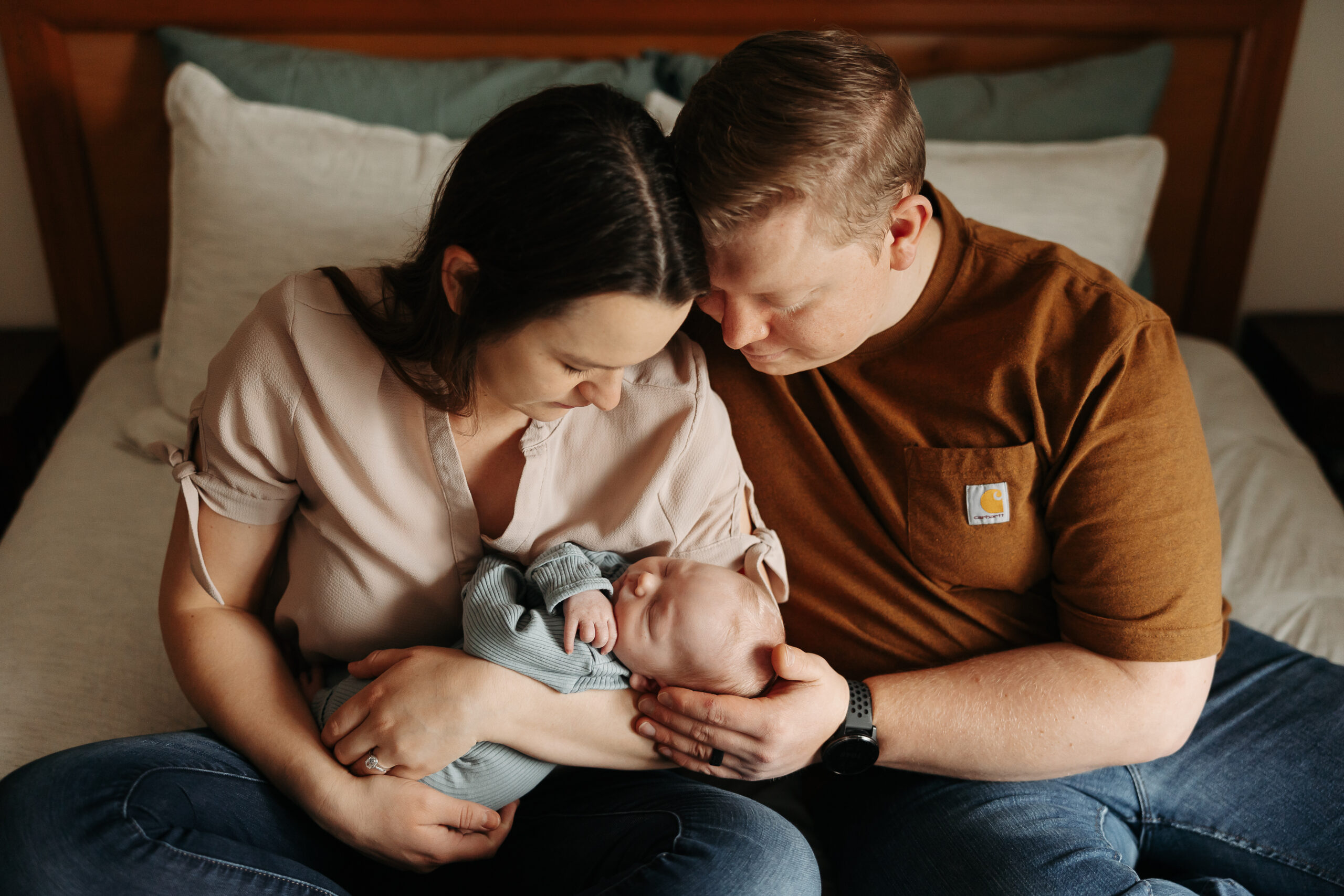 man and woman sitting on bed holding their newborn baby in between them and looking down at him